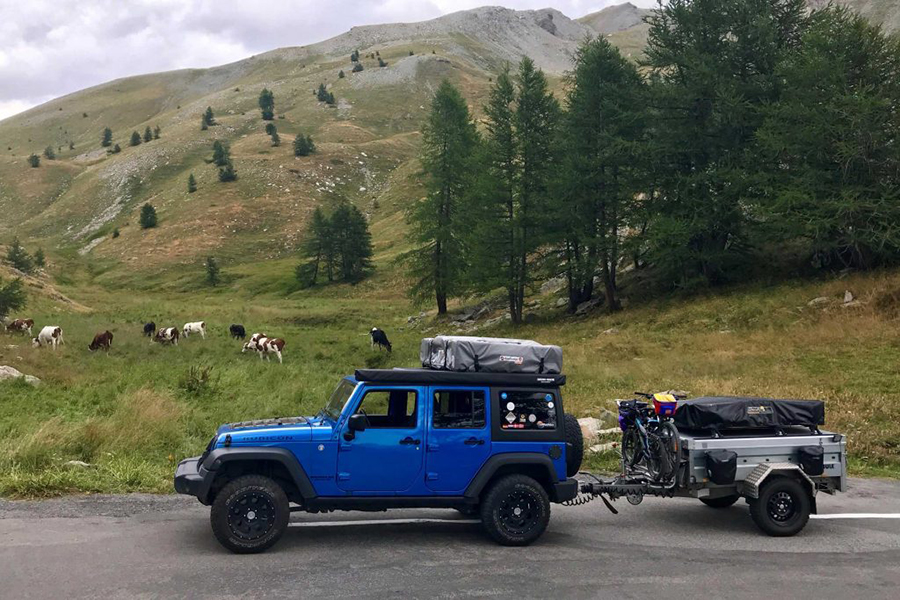 Blauer Geländewagen mit Dachzelt und Anhänger fährt durch Berglandschaft