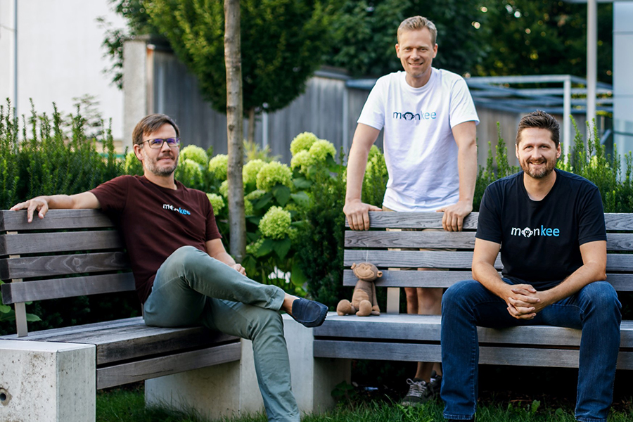 Gruppenbild der drei Gründer der App Monkee. Zwei sitzen auf Holzbänken, einer steht mittig dahinter