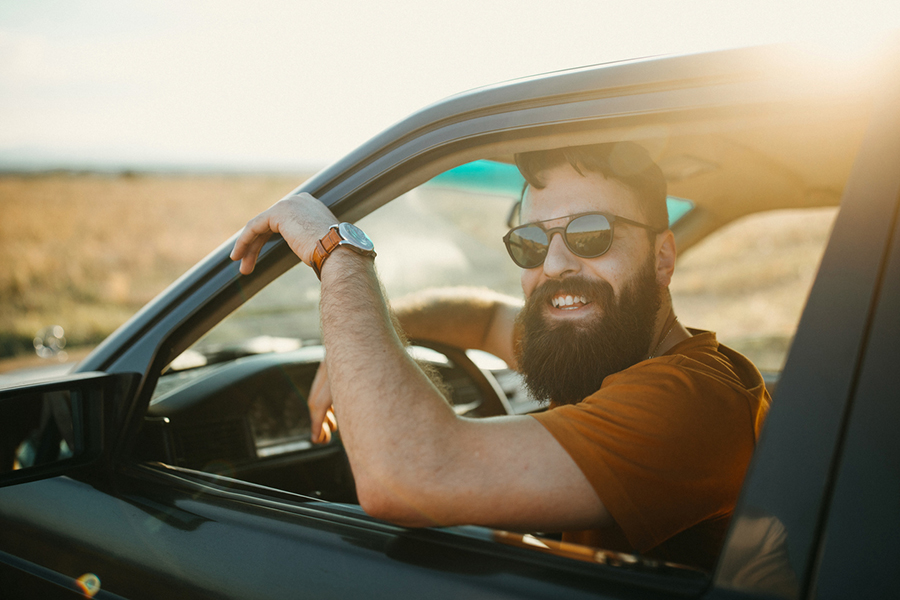 Mann mit Bart und Sonnebrille sitzt am Steuer eines Autos und lächelt in die Kamera