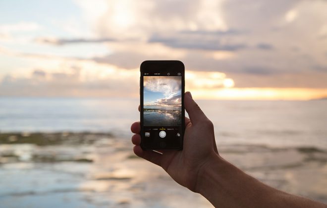 Hand mit Smartphone vor einem Sonnenuntergang hinter dem Meer