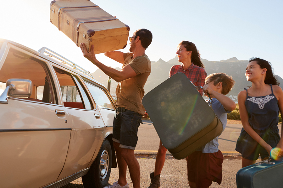 Familie mit Koffern, Mann hebt Koffer aufs Autodach